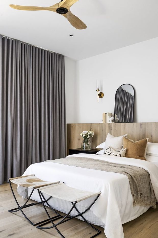A bright, refined bedroom with a white upholstered bed dressed in layered neutral bedding, a grey throw, and assorted beige and white cushions. A full-width timber headboard incorporates a slim black bedside table and styling, with an arched black-framed mirror above. Floor-to-ceiling grey curtains cover the left wall, while a timber three-blade ceiling fan, brass wall sconce, light timber flooring, and two black-framed upholstered benches complete the room.