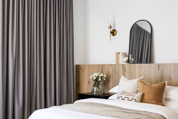 A softly styled bedroom featuring a large upholstered bed with layered white and beige bedding, a warm timber headboard wall, full-height taupe curtains, and a black bedside table. An arched black-framed mirror and brass wall sconce add refined accents, while a small vase of flowers and neutral cushions create a calm, contemporary atmosphere.