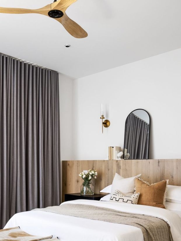 A softly styled modern bedroom with a white ceiling and walls, full-height charcoal-grey curtains, and a timber half-height feature wall behind the bed. The bed is dressed in white linen with neutral beige and tan cushions and a textured throw. A black bedside table, arched black-framed mirror, brass wall sconce, timber ceiling fan, and small floral arrangement complete the calm, warm interior.