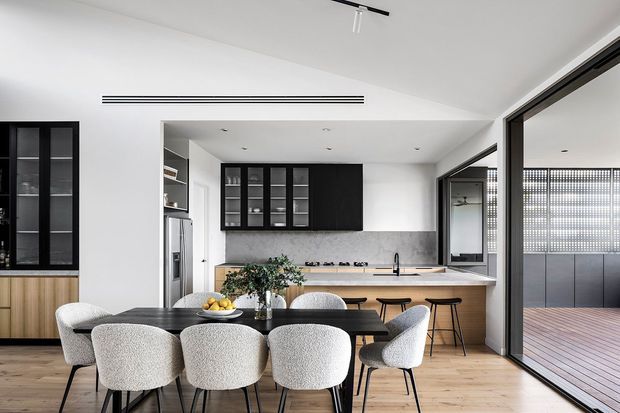 Bright contemporary open-plan kitchen and dining area with white walls, pale timber flooring, black-framed glazing, and integrated dark cabinetry. The kitchen includes a long timber island with a light stone benchtop, black tapware, and bar seating, while a black dining table with upholstered chairs occupies the foreground. Large sliding glazing opens to a timber deck, and a partially visible outdoor area appears beyond.