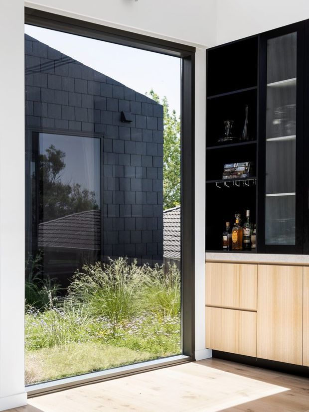 Bright contemporary interior corner with a full-height dark-framed window overlooking a landscaped courtyard and dark block-clad wall. A built-in bar or display cabinet occupies the right side, combining black open shelving, ribbed translucent glass doors, integrated glassware and bottles, and light timber drawers. Pale timber flooring and white walls create a clean, high-contrast composition.