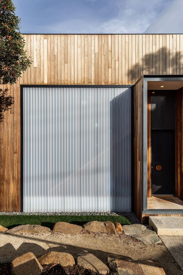 Contemporary residential facade featuring vertical warm timber cladding, a translucent corrugated panel, and a recessed dark front entry framed in timber and metal. A simple stone-edged garden and concrete path occupy the foreground under bright daylight.