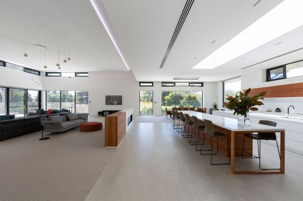 Bright, open-plan contemporary home interior combining a living room, dining area and kitchen. The space features a large kitchen island with extensive white cabinetry, a timber island base, integrated appliances, bar seating, a long dining table, grey sectional seating, pale flooring and broad glazed openings. Linear recessed lighting, concealed air-conditioning slots and skylight-like ceiling openings create a clean, refined atmosphere.