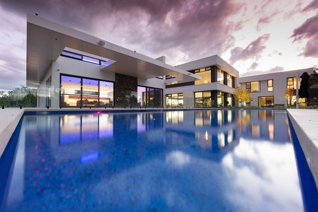A contemporary multi-wing residence viewed from the edge of a large reflecting swimming pool. The home features crisp white and grey geometric volumes, extensive floor-to-ceiling glazing, dark stone cladding, broad flat roof planes, deep overhangs, and a dramatic evening sky reflected in the water. Warm interior lighting contrasts with the cool blue pool and twilight exterior.