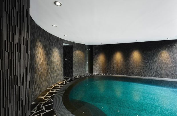 Indoor swimming pool with a curved perimeter, dark textured vertical wall cladding, recessed ceiling lights, and warm wall-washing illumination. The pool is bordered by dark patterned paving and a narrow drainage channel, creating a dramatic, luxurious atmosphere.