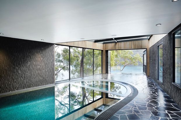 A contemporary indoor pool area with a curved turquoise swimming pool, expansive floor-to-ceiling glazing, dark textured wall cladding, light timber ceiling accents, and irregular dark stone flooring. The glazing reflects surrounding trees and reveals a serene water view beyond.