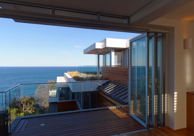 Contemporary coastal residential view from an interior, looking through fully opened floor-to-ceiling glazed doors onto layered timber decks and terraces. The home steps down toward the ocean, with glass balustrades, dark metal roofing, white rendered volumes, timber cladding, and expansive blue-water views.