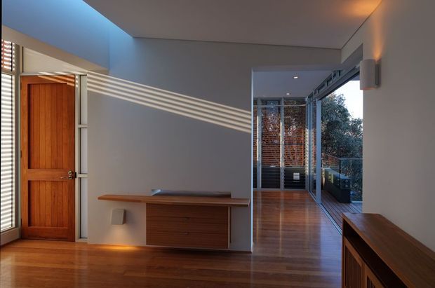 A refined residential entry hall with warm timber flooring, a solid timber front door, and a low built-in timber console with drawers. Strong linear sunlight enters through the entry glazing, creating striped shadows across the pale walls. A glazed corridor extends toward a leafy exterior, with additional timber joinery in the foreground. The space has clean modern lines and a warm, understated palette.