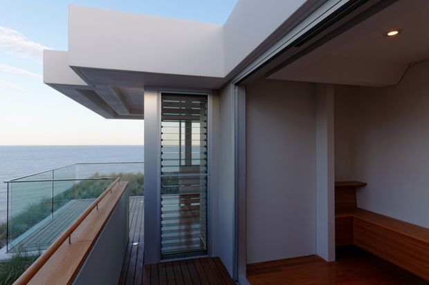 Contemporary coastal residential exterior/interior threshold with a covered balcony or terrace overlooking the ocean. The image shows crisp white rendered walls, projecting flat roof forms, extensive dark-framed glazing, a glass balustrade, timber decking and timber built-in bench seating within the open room.