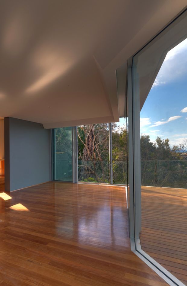 A bright, mostly empty contemporary interior with polished warm timber flooring, a smooth white ceiling, and extensive floor-to-ceiling glazing. The glazing opens visually onto a timber deck and leafy vegetation, with a blue sky beyond. Slim dark window and door frames create a strong vertical rhythm, while a deep projecting ceiling or roof overhang shades the glass.