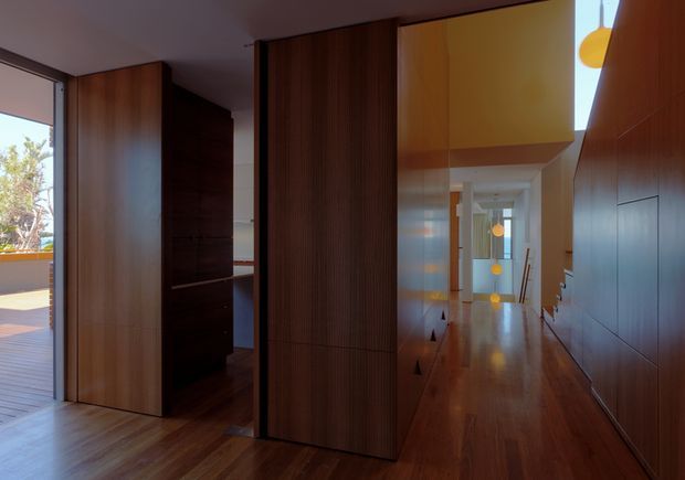 Interior view of a refined contemporary hallway or circulation space with extensive dark timber wall panelling and built-in joinery. A tall central timber screen or pivot door divides the space, while a long polished timber floor leads toward a brighter rear area. Warm yellow architectural volumes and small pendant lights add contrast, with glazing at the left opening toward an exterior terrace or garden.