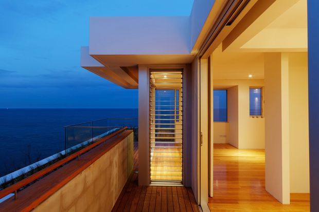 A dusk view from a modern coastal residence, looking along a narrow timber-clad balcony beside open floor-to-ceiling glazing. The interior beyond contains warm timber flooring, white walls, recessed lighting, and a tall louvred or slatted window screen. A frameless glass balustrade and projecting flat-roofed building forms overlook the dark ocean.
