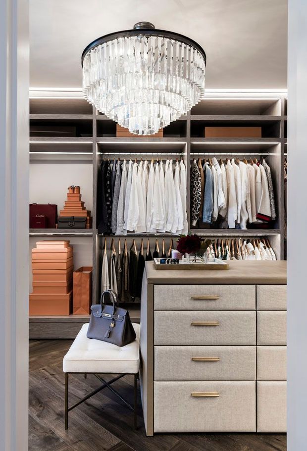 A luxurious walk-in wardrobe with open dark joinery, illuminated clothing rails, neatly arranged garments and stacked boxes. A central upholstered island with multiple drawers occupies the foreground, paired with a bench and handbag. A large tiered crystal chandelier creates a glamorous focal point, while dark timber flooring and warm integrated lighting add contrast.