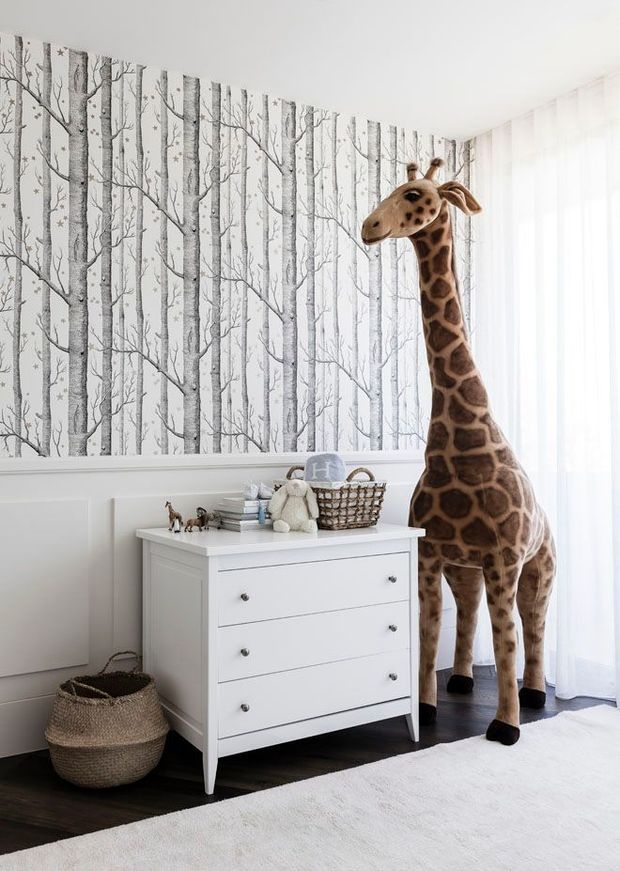 Bright children's bedroom or nursery corner with white painted panelled walls, grey-and-white birch-tree wallpaper, sheer curtains, dark timber flooring and a white four-drawer dresser. A large decorative giraffe stands beside the dresser, with baskets, books and plush toys adding a playful nursery atmosphere.