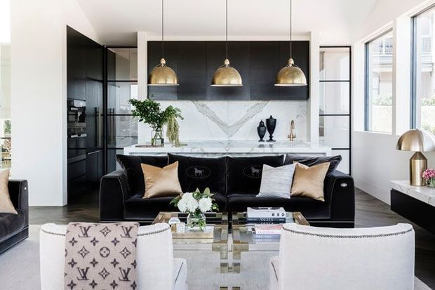 Bright, high-end open-plan interior combining a black kitchen with marble backsplash and island behind a black leather sofa, with white upholstered armchairs in the foreground. Brass dome pendant lights, large windows, dark cabinetry, and polished surfaces create a refined contemporary living and entertaining space.
