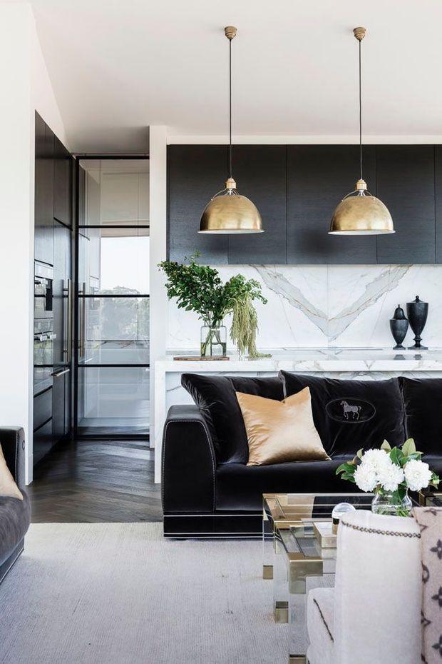 A sophisticated open-plan kitchen and living area with dark flat-panel cabinetry, a dramatic white marble-look splashback, and a white island or benchtop. Two brass dome pendant lights hang above the island. In the foreground is a black leather sofa with a gold cushion, paired with a mirrored glass coffee table and pale upholstered seating. Black-framed glazed doors lead toward a secondary space, while dark timber flooring and a light rug create contrast.