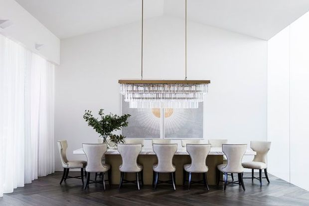 Bright, refined dining room with a long dining table surrounded by upholstered chairs, dramatic twin pendant chandelier, sheer floor-to-ceiling curtains, white walls, dark timber flooring, and a large abstract artwork. The space has a clean, contemporary interior with strong symmetry and abundant natural light.