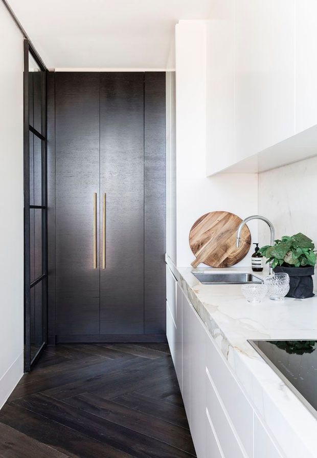 A narrow, refined kitchen with a compact white stone countertop, integrated cooktop, undermount sink, pale marble-look splashback, and white cabinetry. A tall dark timber storage unit with long brass handles anchors the rear, while black-framed glazing and dark herringbone timber flooring add contrast. A small potted plant and timber boards provide natural accents.