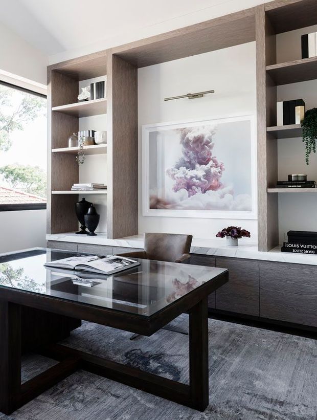 A refined home office with a dark wood desk featuring a glass top, positioned in front of custom built-in shelving and cabinetry. The shelving combines dark timber-look panels with white niches and displays books, ceramics and decorative objects. A framed floral artwork hangs above the low cabinetry, while a large window provides abundant natural light. The room has a sophisticated neutral palette with grey carpeting and dark wood furniture.