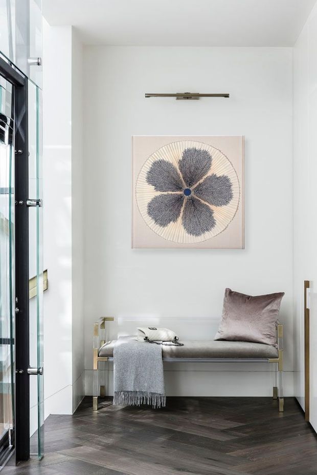 A bright, refined entryway or hallway with white walls, dark herringbone timber flooring, and a minimalist upholstered bench with clear acrylic or glass side supports and metallic legs. A large floral artwork and slim wall light create a subtle focal point, while a tall glass-fronted cabinet or doorway is visible along the left side.