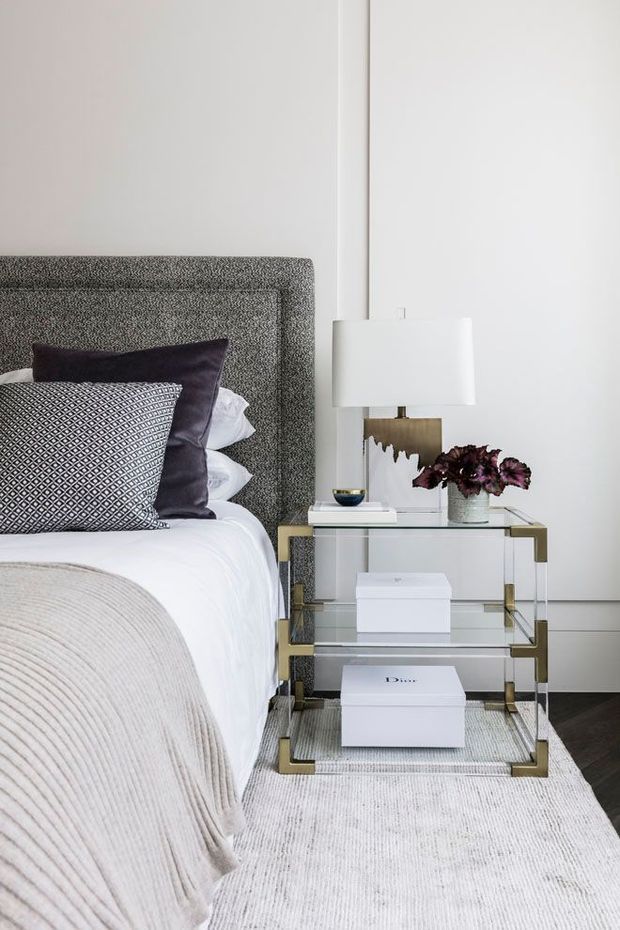 A refined bedroom vignette featuring a textured grey upholstered headboard, white bedding, layered charcoal and patterned pillows, and a soft beige throw. A glass-and-brass bedside table holds a white lampshade, decorative objects, and flowers. The room has white walls, dark timber flooring, and a pale textured rug, creating a polished, neutral-toned composition.