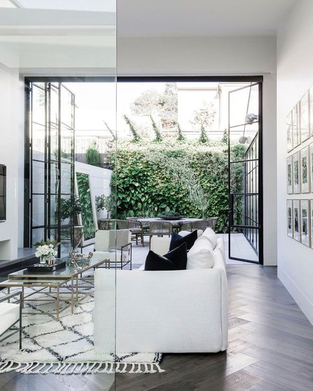 Bright contemporary living room with a white sectional sofa, glass coffee table, patterned rug, framed artwork, and dark herringbone timber flooring. Full-height black-framed glazed doors open onto a lush enclosed courtyard, creating a strong indoor-outdoor connection. The room has abundant natural light and a high, airy feel.