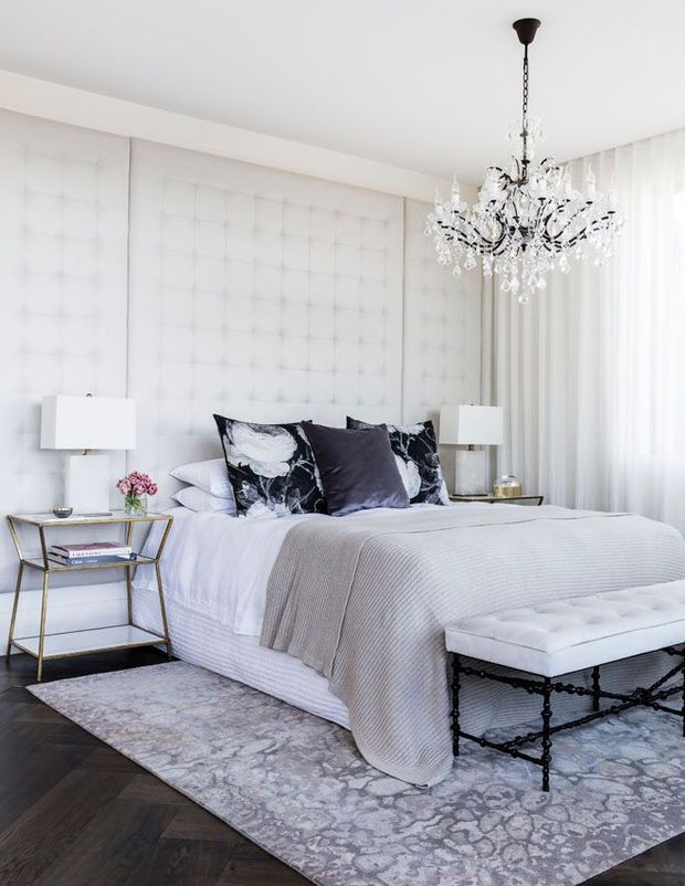 Bright, refined bedroom with a large upholstered, tufted feature wall behind the bed, layered white and grey bedding, patterned accent pillows, and a crystal chandelier. Matching bedside lamps sit on slim gold-toned side tables, while a cushioned black metal bench rests at the foot of the bed. Dark timber flooring is partially covered by a pale patterned rug, and sheer curtains provide soft daylight.