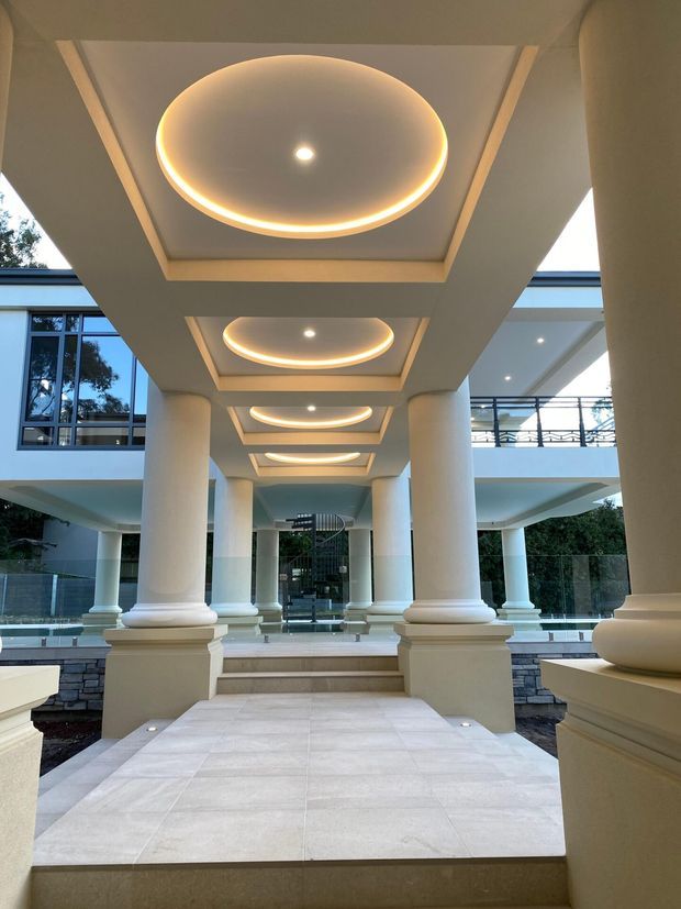 A grand covered exterior entry courtyard or portico with a long colonnade of substantial white classical columns and moulded bases. The tiled walkway leads up shallow steps toward a glazed entrance, while a sculptural coffered ceiling features repeated circular recessed light wells with warm concealed illumination. Modern glazed building wings and an upper-level balcony surround the space, creating a refined transitional classical-contemporary entrance.