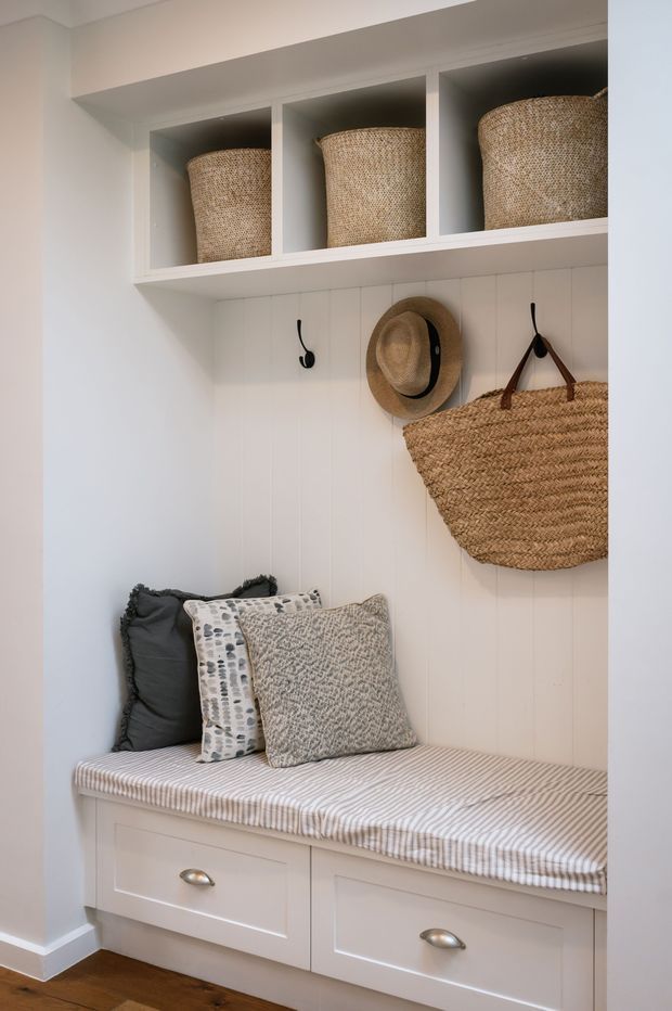 A compact, bright entryway or mudroom nook with white vertical wall panelling, built-in overhead cubby shelving, and a white built-in bench with drawers. Woven baskets, a straw hat, a large woven tote, black hooks, and layered cushions add natural texture. The bench has a soft striped seat cushion and sits above a warm timber floor.