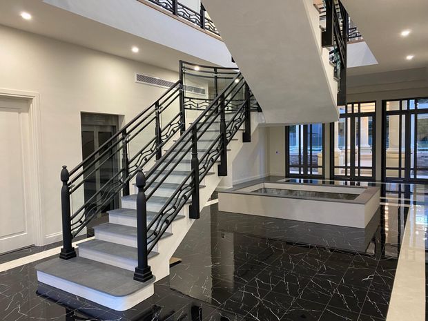 A grand interior foyer dominated by a sweeping staircase with ornate black metal balustrades, white risers, and dark stone treads. The space features glossy black marble-look flooring with light veining, cream walls, recessed ceiling lighting, a large central stair void, upper-level galleries, and full-height glazed doors opening toward an exterior pool or terrace.