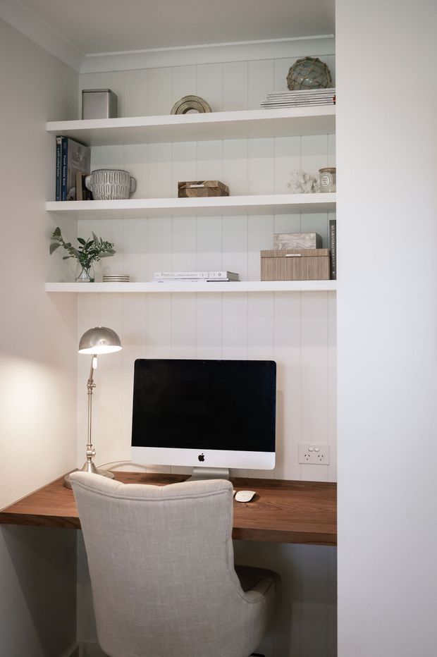 A compact built-in home office nook with a timber desk, desktop computer, upholstered chair, desk lamp, and white open shelving displaying books and decorative objects. The space has white vertical wall panelling, warm timber tones, and soft neutral furnishings.