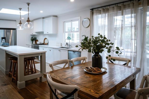 Bright open-plan kitchen and dining area with white shaker-style cabinetry, a large white island with timber bar stools, stainless steel appliances, dark timber flooring, and a substantial rustic timber dining table. Sheer curtains, pendant lights, greenery, and warm natural light create an inviting refined farmhouse-inspired interior.