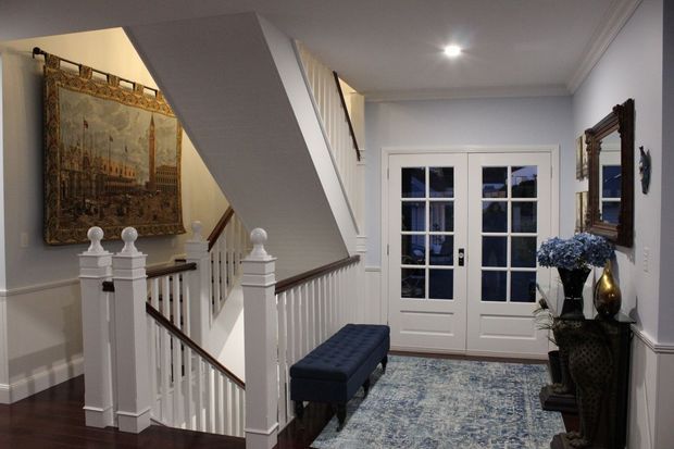 A refined residential entry hall with a staircase, white-painted balustrade and newel posts, dark timber handrails and flooring, pale grey walls with traditional trim, French doors, a blue upholstered bench, patterned area rug, decorative mirror, console table, and wall artwork. The space has a formal, classic character with balanced lighting and cool blue-grey tones.