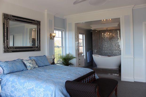 A refined bedroom with a blue patterned bedspread, upholstered bench, ornate framed mirror, pale blue-gray walls, and traditional white wall mouldings. Through the open doorway is a luxurious bathroom featuring a freestanding white bathtub, chandelier, and dark mosaic-tiled feature wall.