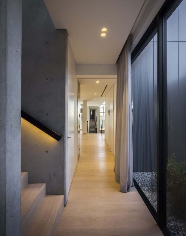 A contemporary interior hallway with pale timber flooring, white walls and ceiling, and a striking exposed concrete stair enclosure on the left. A black handrail with concealed warm lighting follows the stair, while full-height dark-framed glazing and sheer curtains line the right side, looking onto a narrow gravel courtyard. Recessed ceiling lights and a distant stairway reinforce the clean, minimalist architectural character.