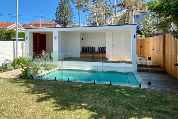 A bright contemporary backyard featuring a compact white pool house or pavilion beside a turquoise swimming pool. The structure has broad open glazing, a covered lounge area with built-in timber seating, a dark timber entry door, white walls, and a timber-decked side path enclosed by a vertical timber fence. A frameless glass pool barrier separates the pool from the lawn, with established trees and neighbouring homes beyond.