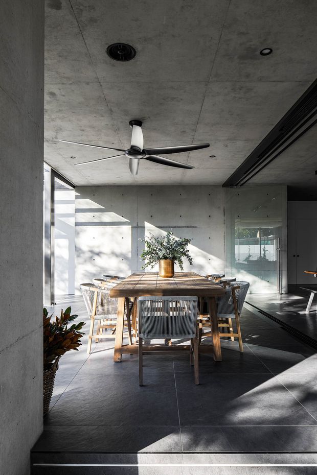 A contemporary dining area with a large rectangular timber dining table and woven dining chairs, set within a minimalist concrete interior. The space features exposed concrete walls and ceiling, dark tiled flooring, expansive glazing, a sculptural ceiling fan, and strong natural sunlight creating patterned shadows. A partially visible kitchen or living area appears beyond the glass partition.