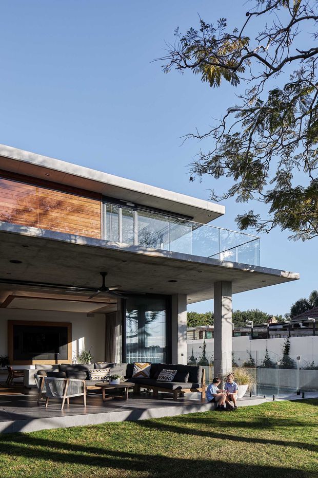 Contemporary two-storey home photographed from the rear garden, featuring a broad cantilevered concrete balcony, warm horizontal timber cladding, extensive glazing, glass balustrades, and an open covered outdoor living area with lounge seating. A landscaped lawn and rear pool area are visible, with mature trees framing the composition.