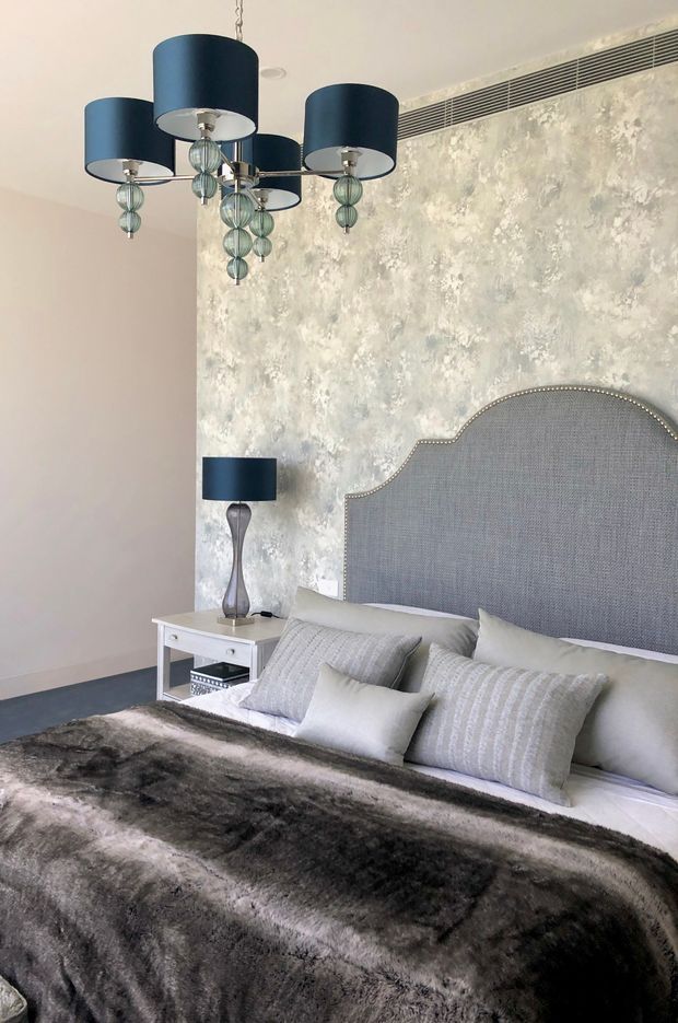 A refined bedroom interior featuring a large upholstered bed with a scalloped, nailhead-trimmed grey headboard, layered pale grey and white bedding, and a dark faux-fur throw. A sculptural multi-arm pendant with navy drum shades hangs over the room, complemented by a matching bedside lamp. The walls include a textured grey-white feature finish and a linear air-conditioning grille, while the flooring appears dark grey carpet.