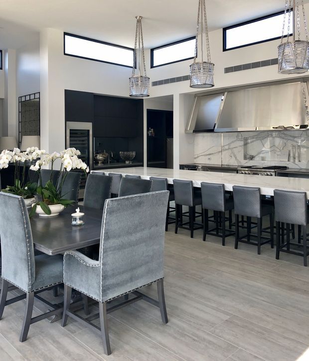 A sophisticated open-plan dining and kitchen interior. The foreground features a large dark dining table with upholstered grey dining chairs, while the rear has a long white kitchen island with dark grey bar stools. The kitchen includes dark built-in cabinetry, a marble-look splashback and benchtop, a large stainless-steel rangehood, and sculptural pendant lights. High clerestory windows bring in natural light, creating a refined contemporary atmosphere.