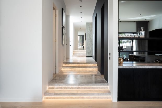 A contemporary interior hallway with a sequence of broad steps leading toward a brighter corridor and mirror at the rear. The hallway has pale walls, polished light stone flooring and stair treads with integrated warm LED strip lighting. Dark vertical timber or charcoal-toned cabinetry and wall panels line the right side, opening to a sophisticated home bar or kitchen area with dark joinery, shelving, glassware and a stone counter. Minimal framed artwork and recessed ceiling lights complete the refined, high-contrast scheme.