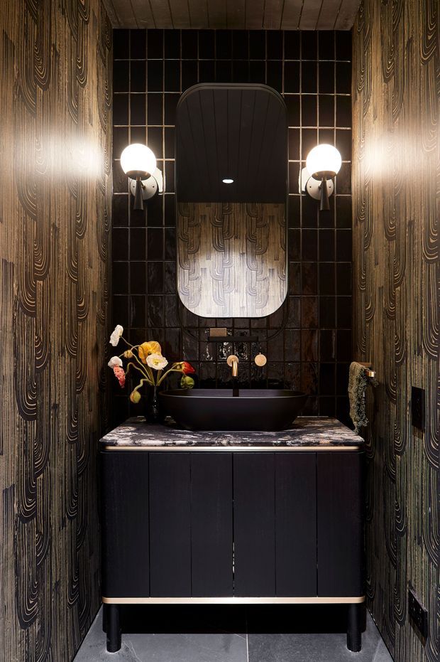 A compact, dramatic powder room featuring a black vessel basin on a dark vanity with a marbled stone benchtop. The walls are clad in ornate dark patterned panels, while the rear wall uses glossy black grid-pattern tiles. A large rounded mirror, sculptural globe wall sconces, brass-toned tapware, and floral arrangements create a moody, luxurious atmosphere.