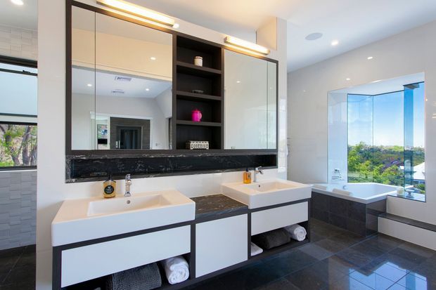 Bright contemporary bathroom with a double vanity, glossy white drawer cabinetry, dark stone benchtops and splashback, large mirrored wall cabinets, and a freestanding bathtub beside floor-to-ceiling glazing. Dark tiled flooring and black-framed glazing add contrast, while integrated lighting creates a clean, polished look.
