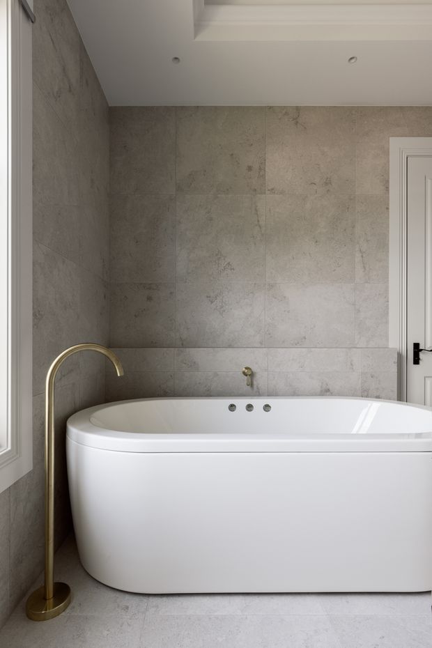 A refined bathroom featuring a freestanding white bathtub set against large grey-beige stone-look wall and floor tiles. A brushed brass floor-mounted curved bath filler stands beside the tub, with white panelled trim, a recessed ceiling detail, and a black door lever adding contrast.