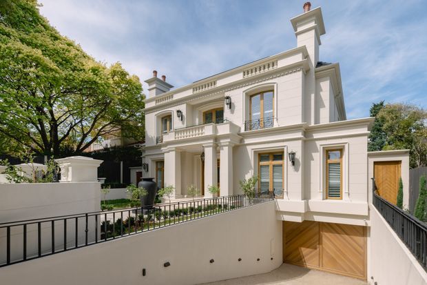Street-facing view of a grand two-storey white-rendered residence with strong classical detailing, including a symmetrical facade, pilasters, decorative balustrades, cornices, tall shuttered windows, columns and a prominent chimney. The home sits above a sloped driveway leading to a timber double garage door, with landscaped trees and black metal railings around the front terrace.