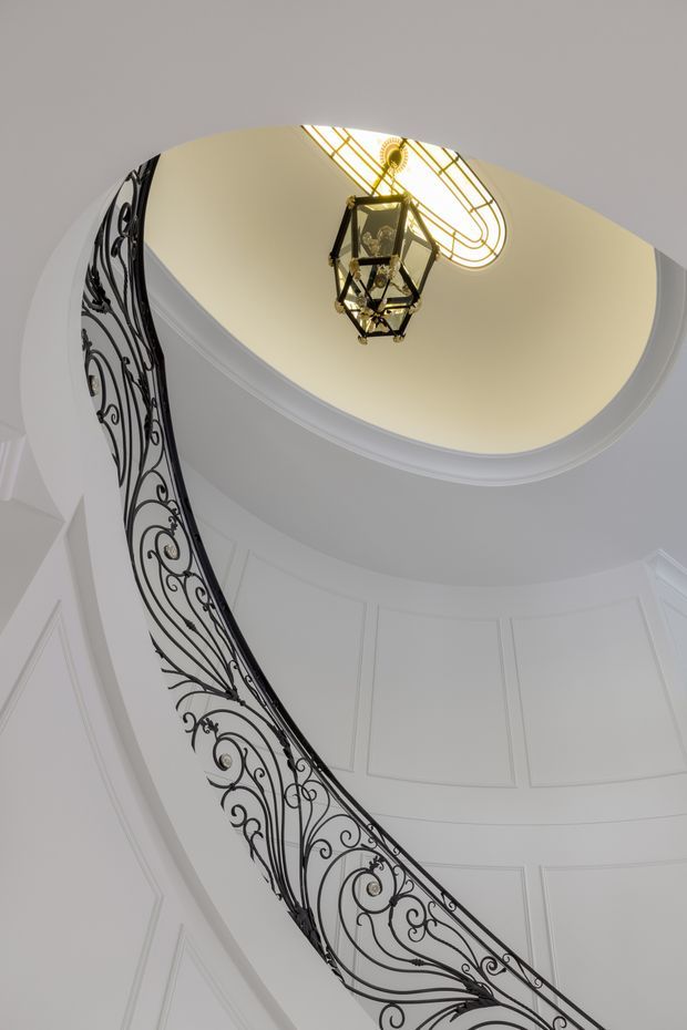 Upward-looking view of an elegant curved stairway or landing beneath an oval domed ceiling. The space features ornate black wrought-iron scrollwork balustrading, white decorative wall panelling and moulded ceiling trim, and a central hanging lantern beneath a glazed skylight or decorative ceiling window. The composition highlights classical detailing and a dramatic vertical void.