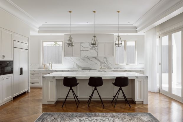 A bright, refined kitchen with a large central island and three dark bar stools. The room features white paneled cabinetry, a dramatic marble backsplash and benchtop, integrated appliances, pendant lighting, timber parquet flooring, and tall glazed doors opening to the outside.
