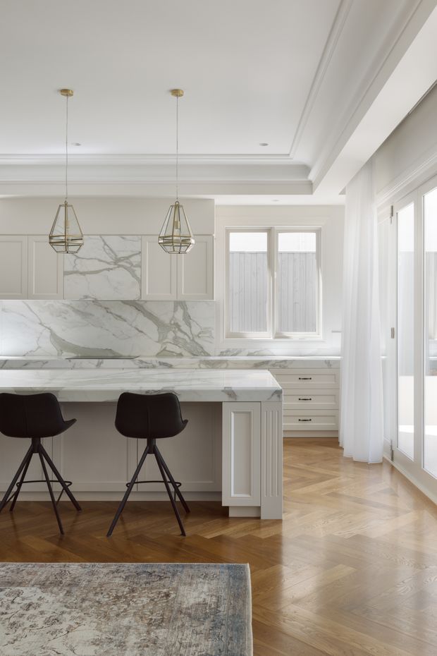 Bright, refined kitchen with a large marble island and full-height marble splashback, pale grey panelled cabinetry, dark bar stools, and brass-toned geometric pendant lights. The room features herringbone timber flooring, layered coffered ceiling trim, large glazed doors and windows with sheer curtains, and a neutral, elegant palette.