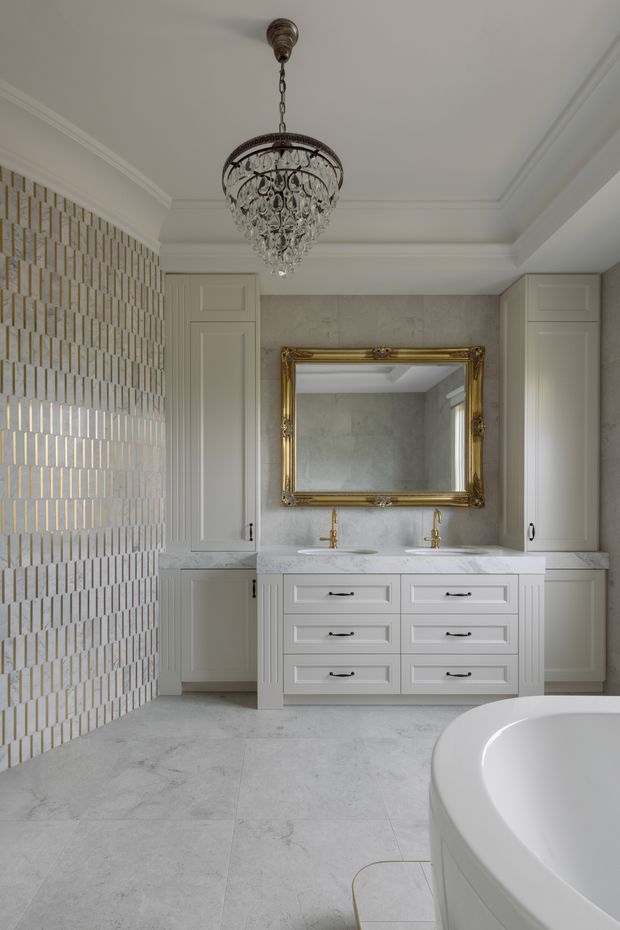 Elegant, traditional bathroom with a large double vanity, white shaker-style cabinetry, twin gold-toned faucets, and a large ornate gilt-framed mirror. The room features pale marble-look floor and wall surfaces, a decorative geometric wall tile with metallic accents, white panelled walls and cabinetry, and a crystal chandelier. A freestanding bathtub is partially visible in the foreground.