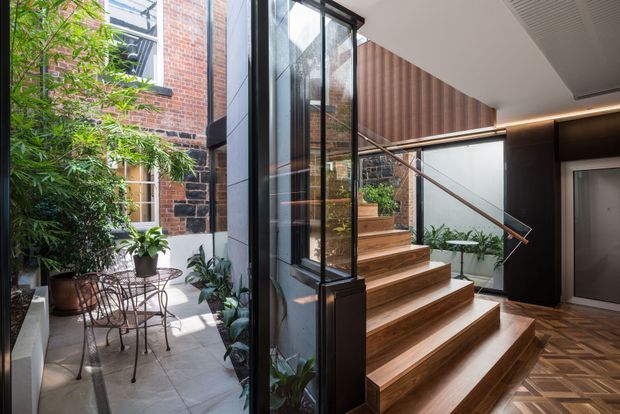 A refined residential entry and stair hall arranged around a narrow landscaped courtyard. Full-height glazing connects the interior to the planted outdoor space and reveals an adjacent brick building. Warm timber stairs with a slim metal handrail rise beside dark built-in wall panelling and vertical timber detailing, while patterned timber flooring and concealed linear lighting add visual warmth.